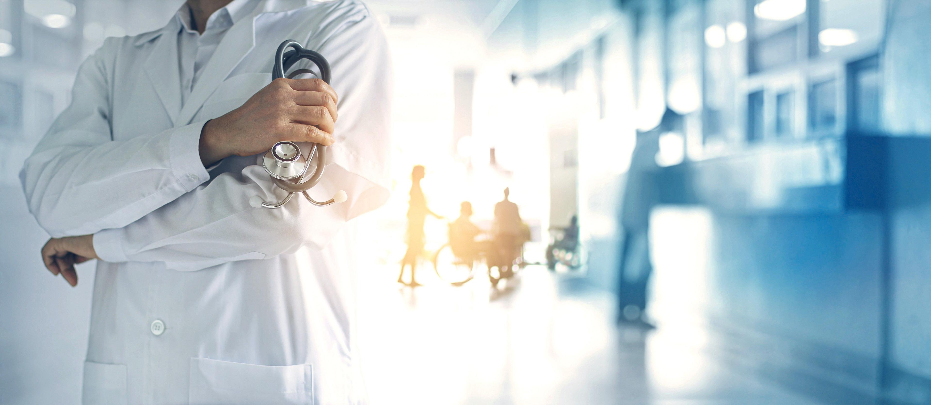 Doctor in white lab coat holding a stethoscope in a bright hospital hallway.