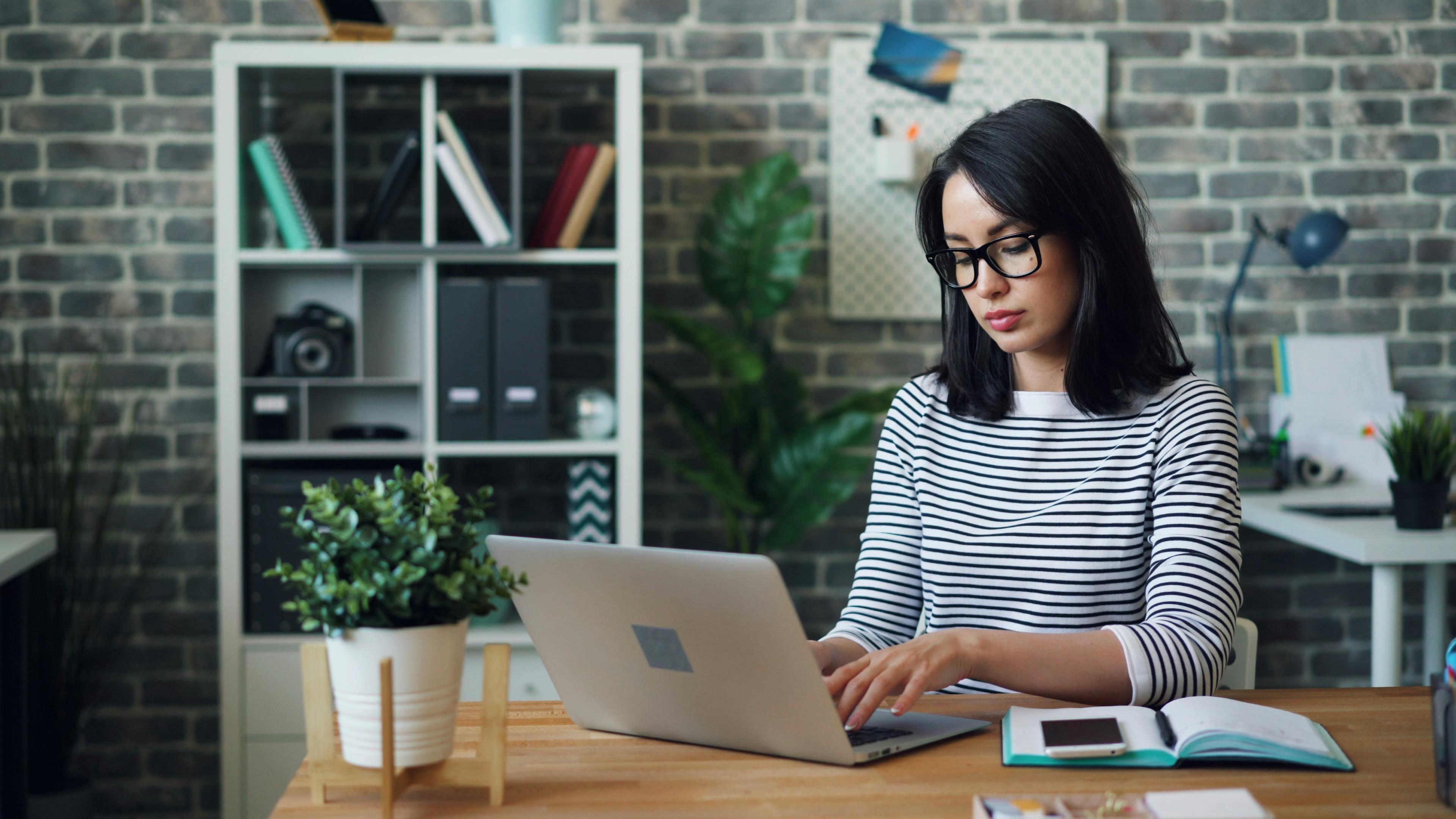 Young woman with glasses typing on a laptop at a wooden desk.
