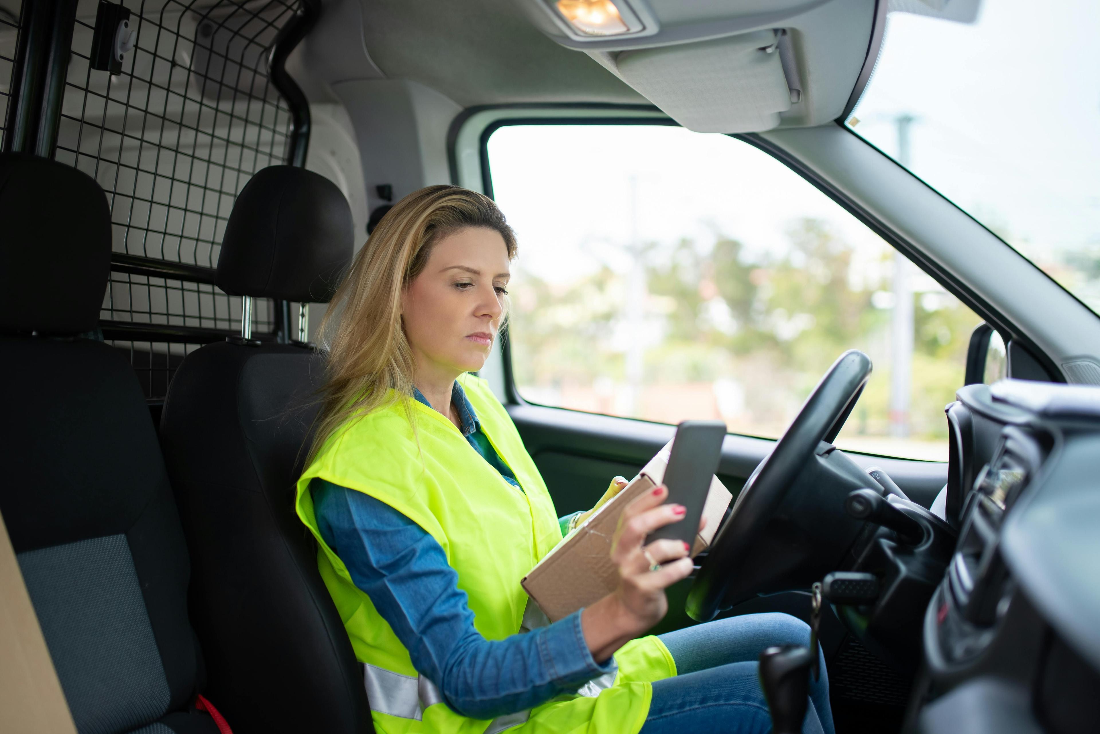 Blonde woman in a high-visibility vest in a van, operating a digital tablet.