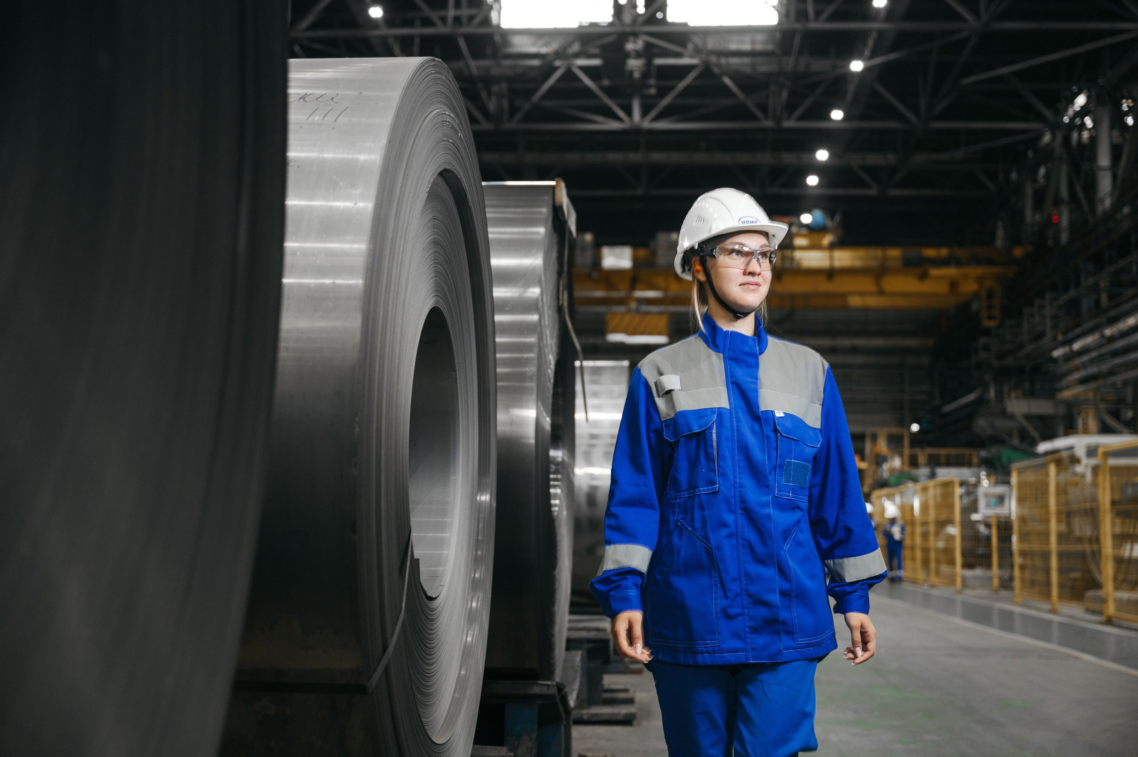 Young man in hard hat and blue overalls walks past large metal coils in a factory.