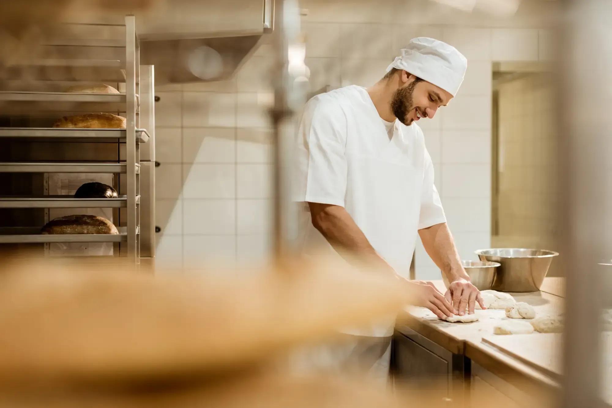 Smiling baker in white uniform kneading dough skillfully in a professional kitchen.