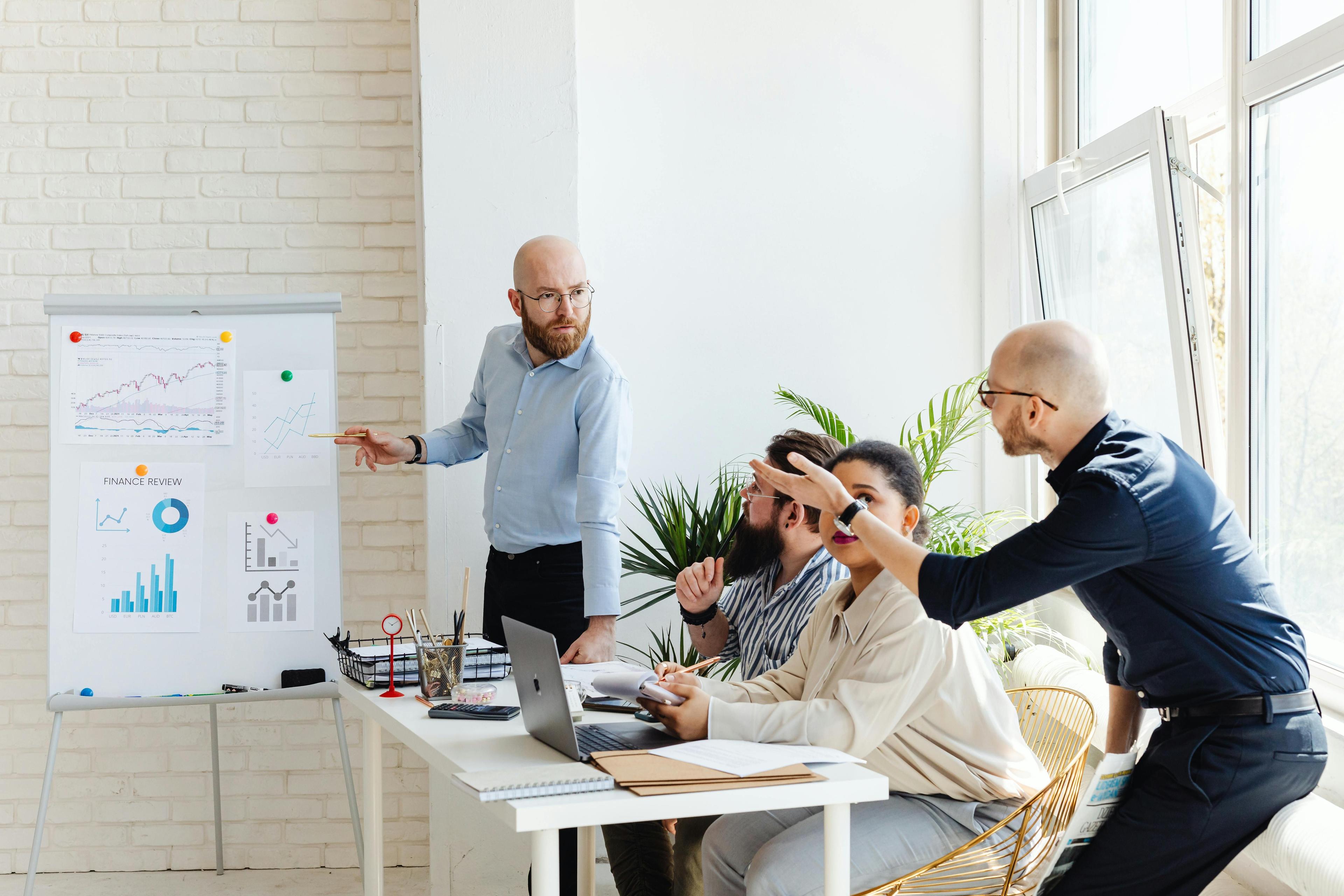 A man presents charts on a whiteboard to three colleagues in a bright meeting room.