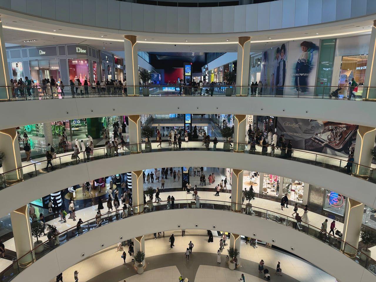 Spacious multi-level shopping mall interior with numerous people browsing shops.