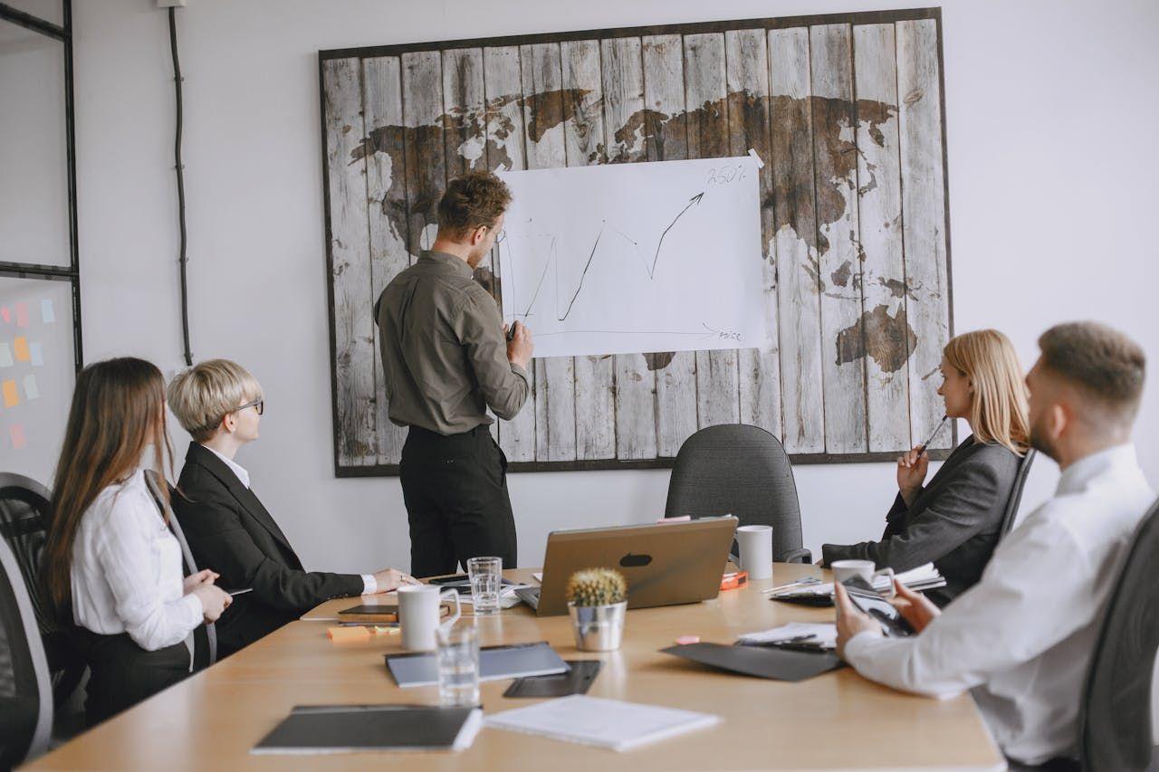 A man presents a graph on a whiteboard to five colleagues in a modern meeting room.