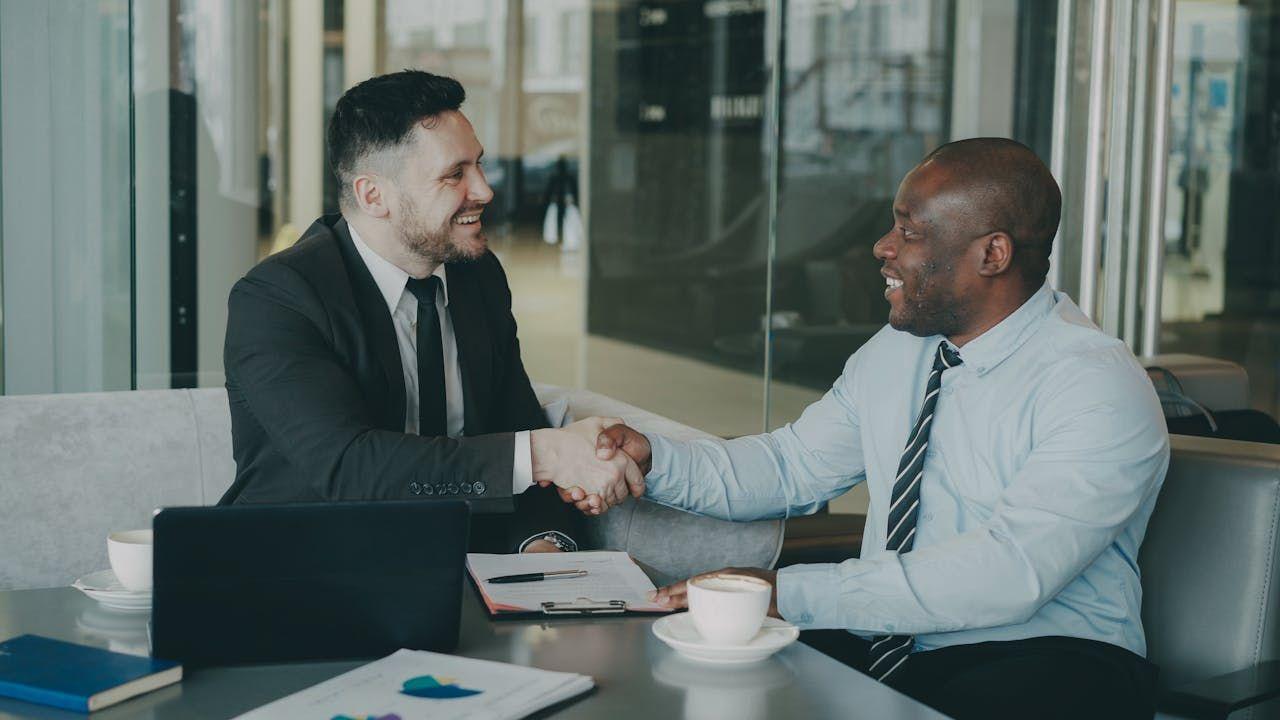 Two diverse businessmen shaking hands over a table with a laptop and coffee.